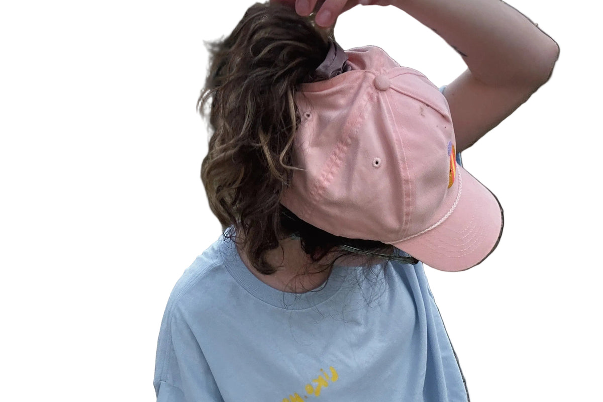 A person with brown hair wearing a light pink baseball cap backwards and a light blue t-shirt, shown against a solid black background.
