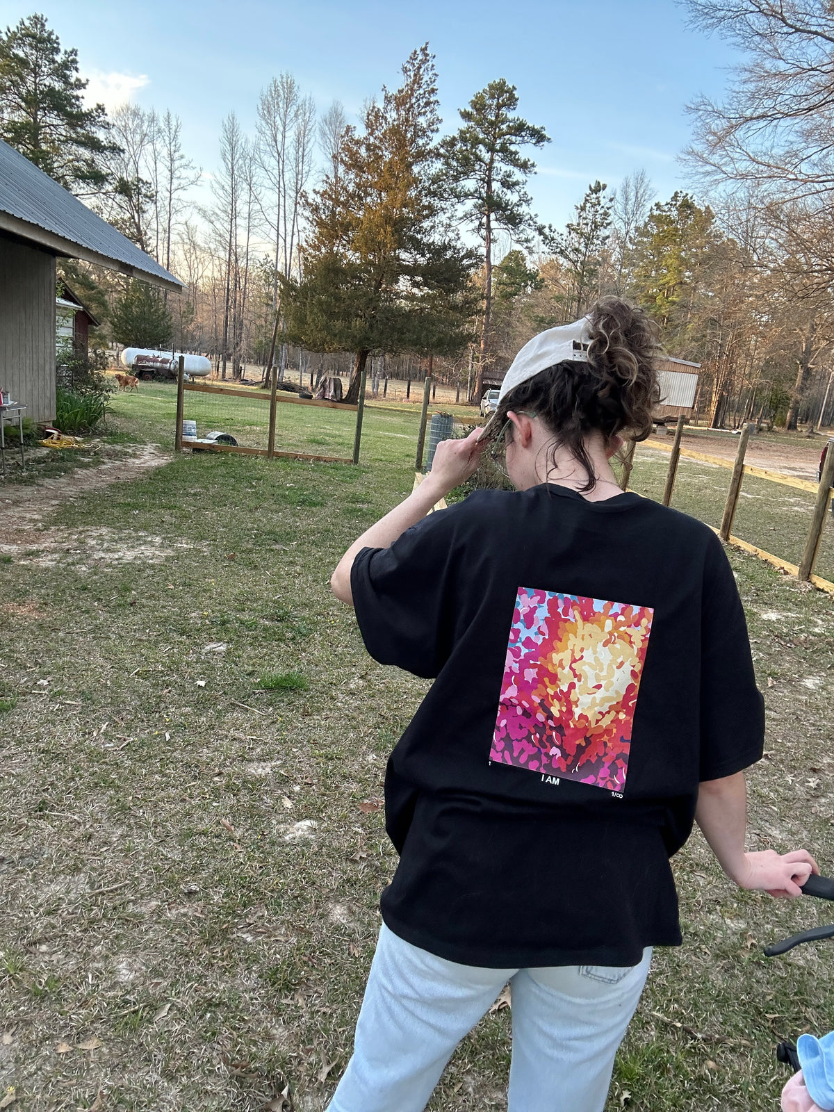 A person standing outdoors in a grassy yard wearing a black oversized T-shirt with a large colorful abstract floral or fire-like graphic on the back, paired with light gray pants and a light-colored cap.